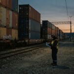 Cargo containers at a border crossing at dusk, representing who benefits from a proxy war through distant trade and supply routes